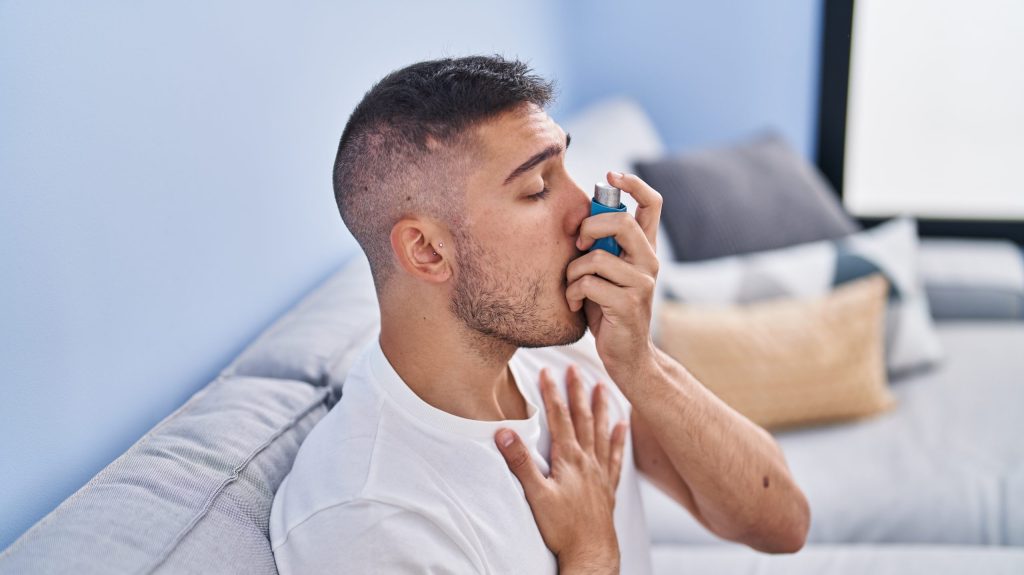 Young hispanic man using inhaler sitting on sofa at home thumbnail